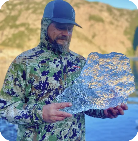 A man in a hooded jacket holding up a catch, water behind him.