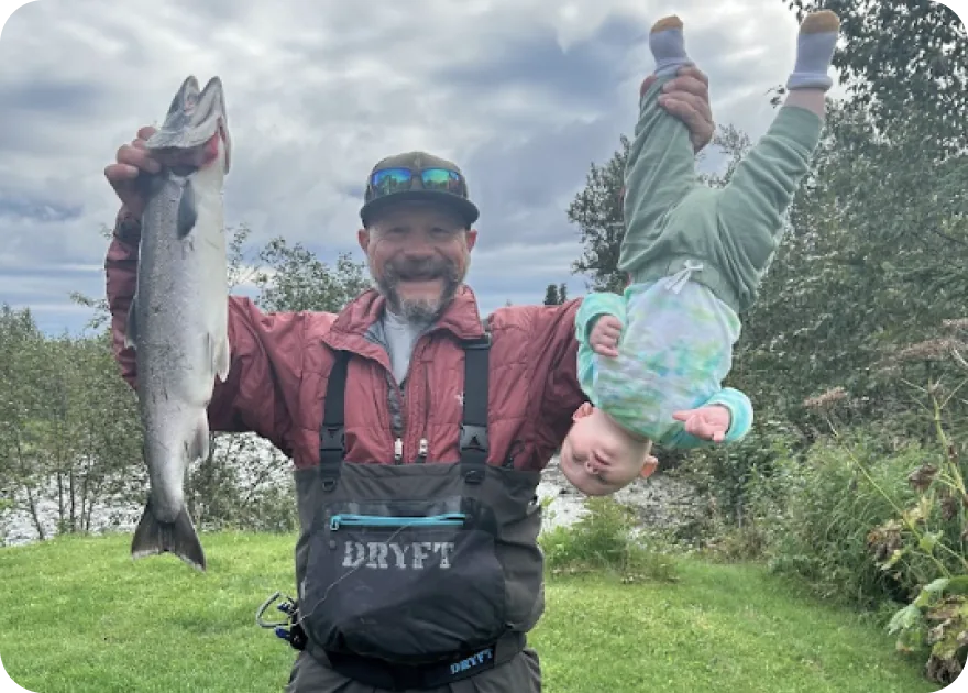 A man in a red jacket holding up two salmon, friends behind him on the bank.