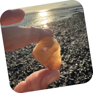 A hand holding a small amber-coloured stone up against the light on a pebble beach.